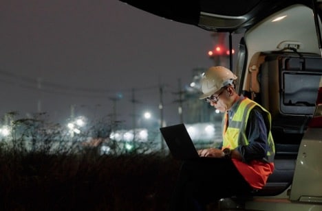 An engineer sits on a piece of heavy machinery, working on a laptop, with a power station in the background.