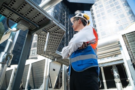 An engineer stands under air conditioning and water lines in a busy city environment to inspect utilities and ensure quality.