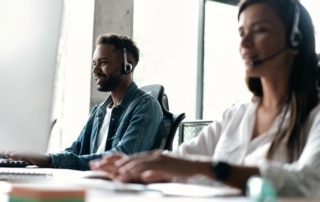 Male and female call center agents sit typing at a desk behind a computer and wearing headsets