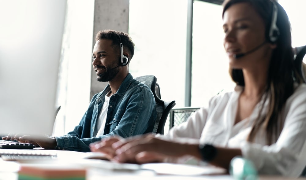 Male and female call center agents sit typing at a desk behind a computer and wearing headsets