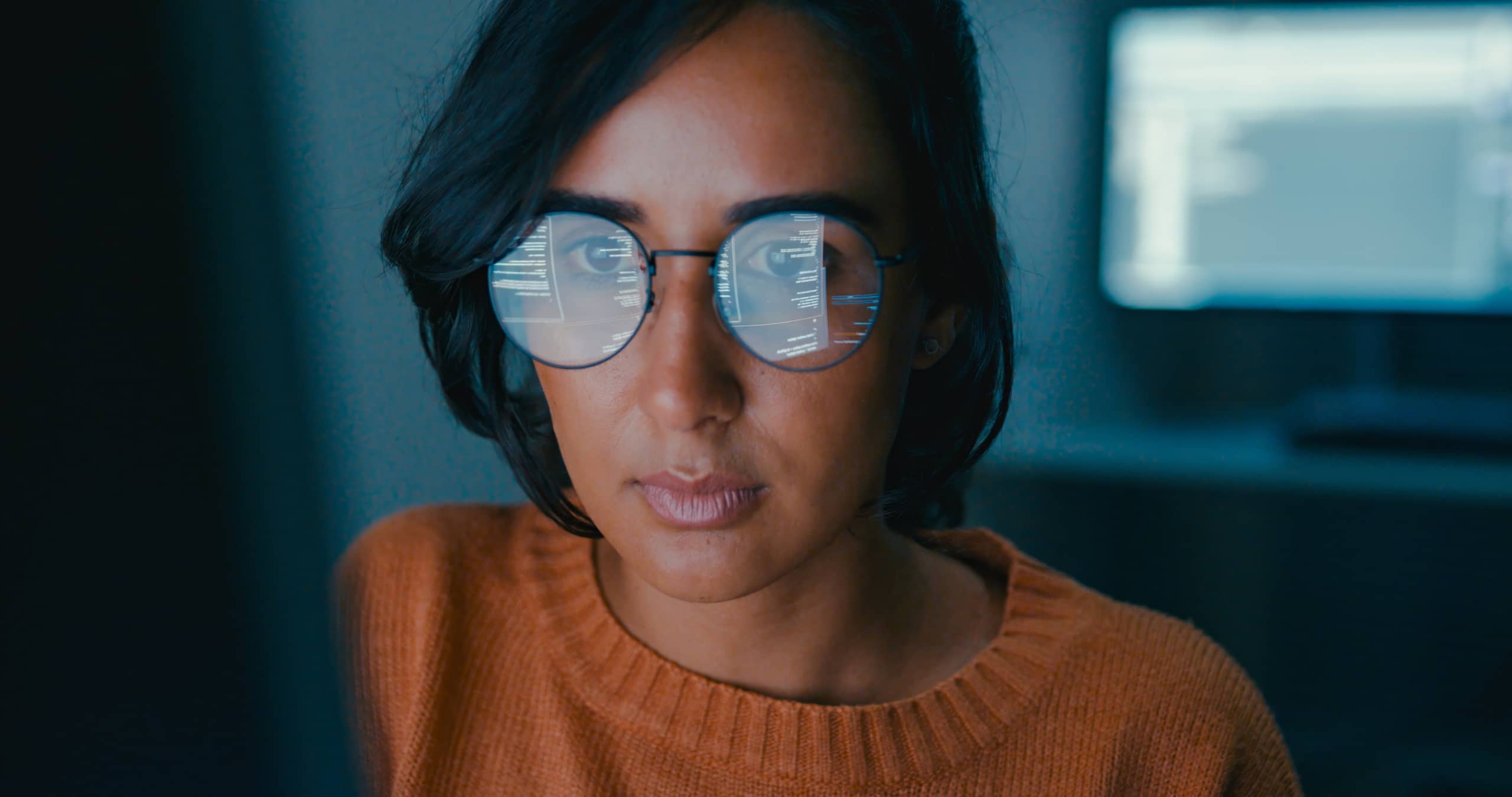  A programmer works diligently on an AI call center tool. She is sitting in a dark room, and her computer monitor is reflected in her glasses.