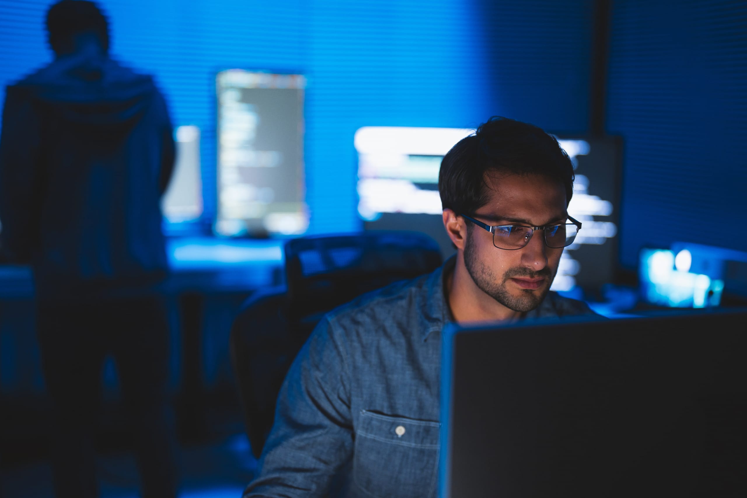 A programmer working at a computer in a dark room, lit by screens covered in code.