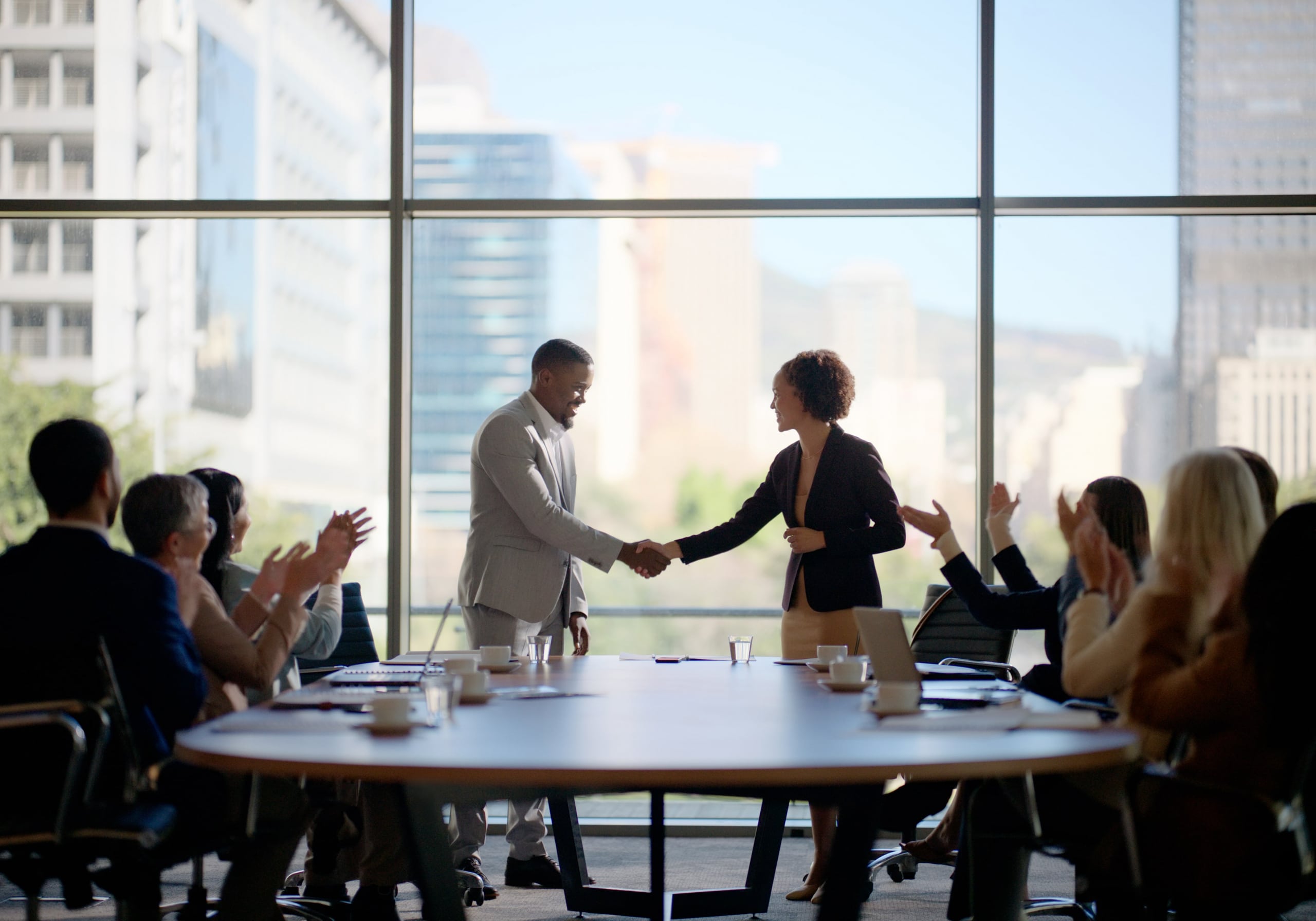 Two teams of professionals on either side of a large table with their leaders up front, shaking hands.