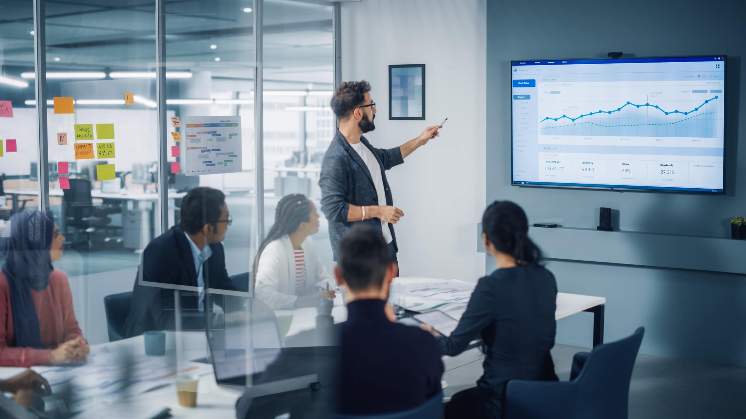 A group of professionals sitting at a desk in a meeting led by a male pointing at a screen with a chart.