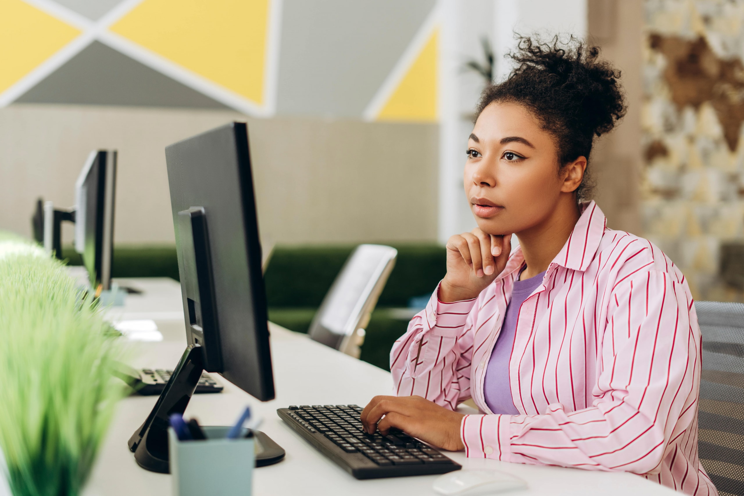 Young office worker is concentrating on her computer screen, typing on the keyboard and thoughtfully resting her chin on her hand, showcasing a productive work environment