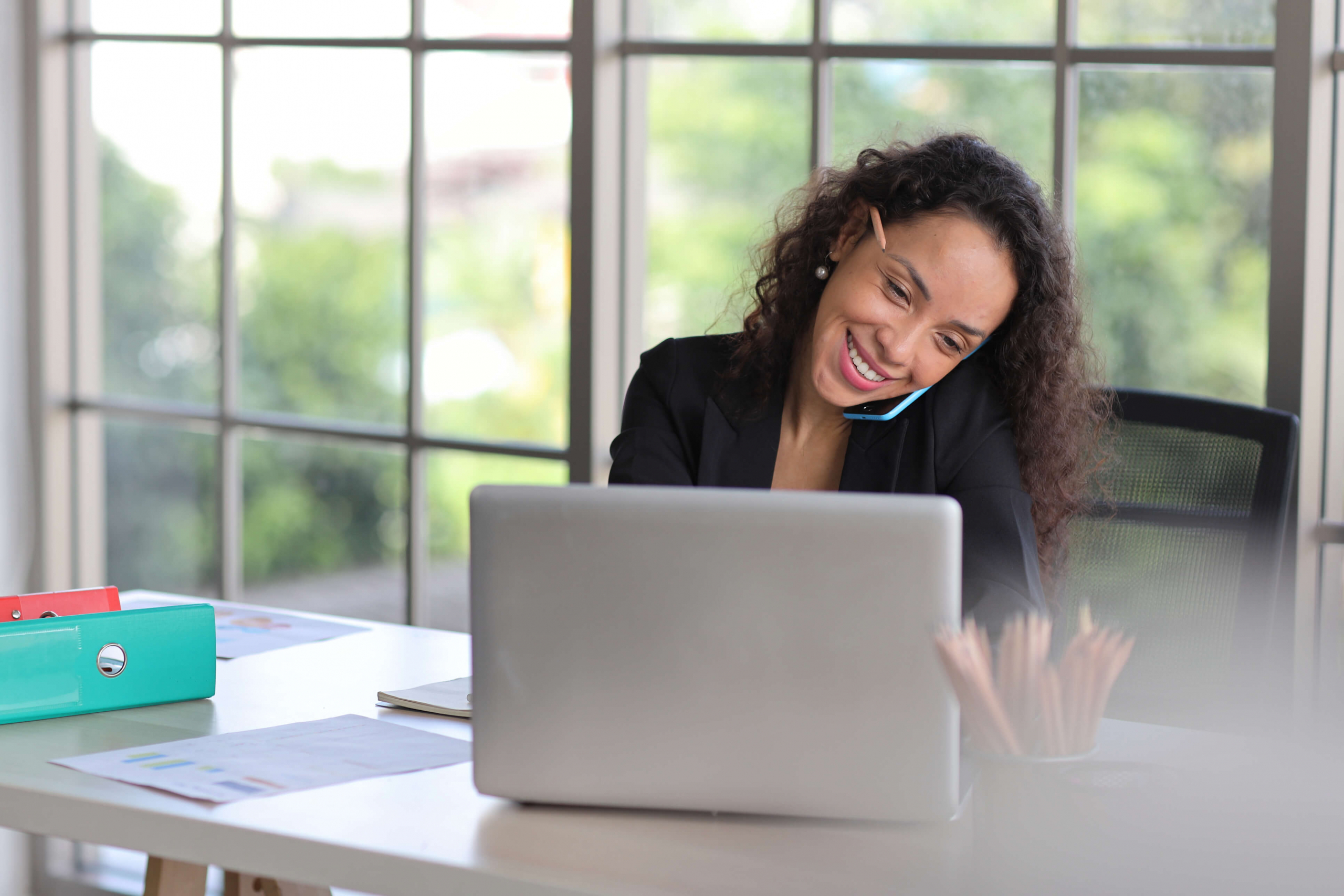 Woman talking on her cell phone smiling while sitting and looking at her laptop