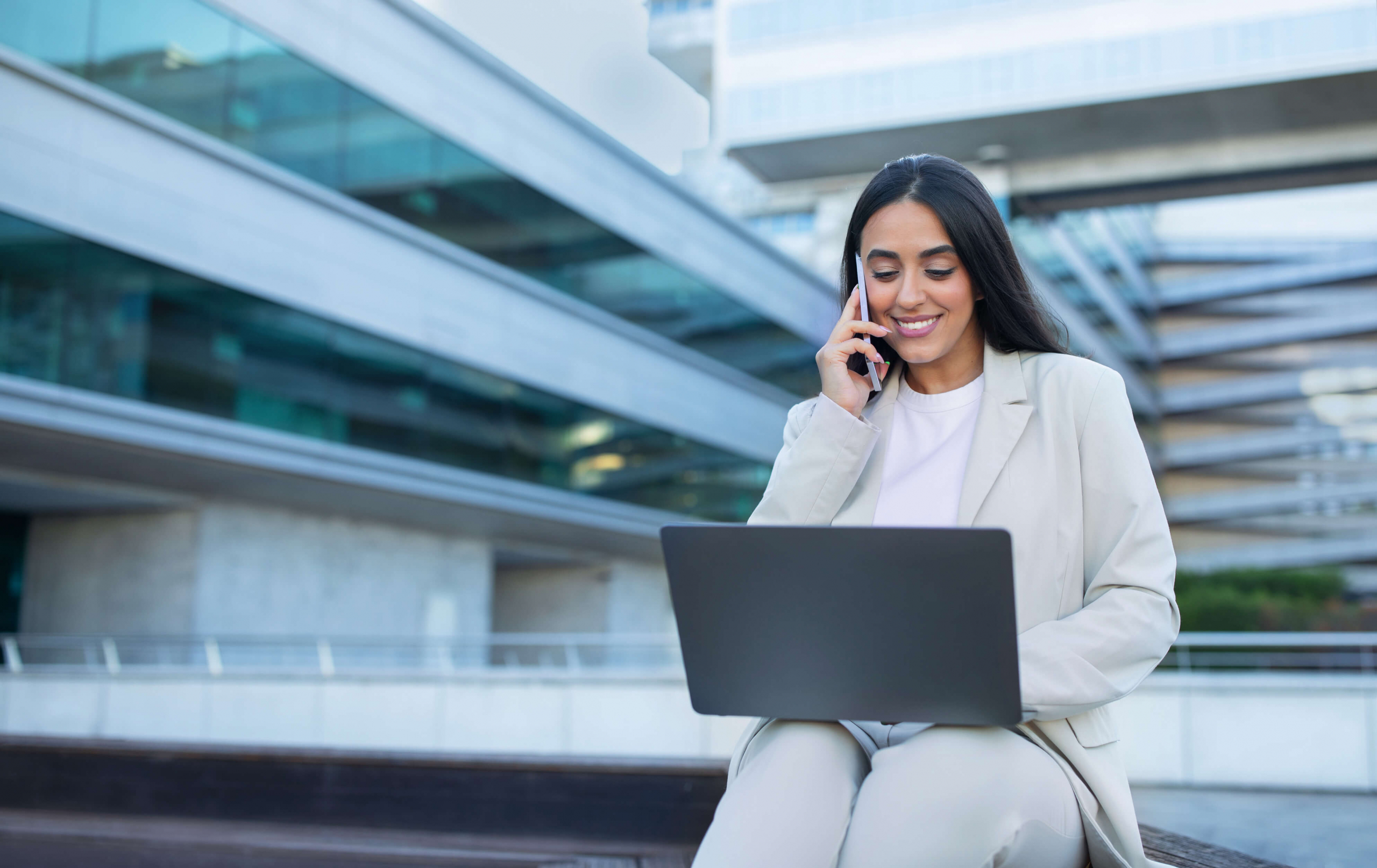 4. Woman smiling on her phone and holding her laptop sitting outside an office building