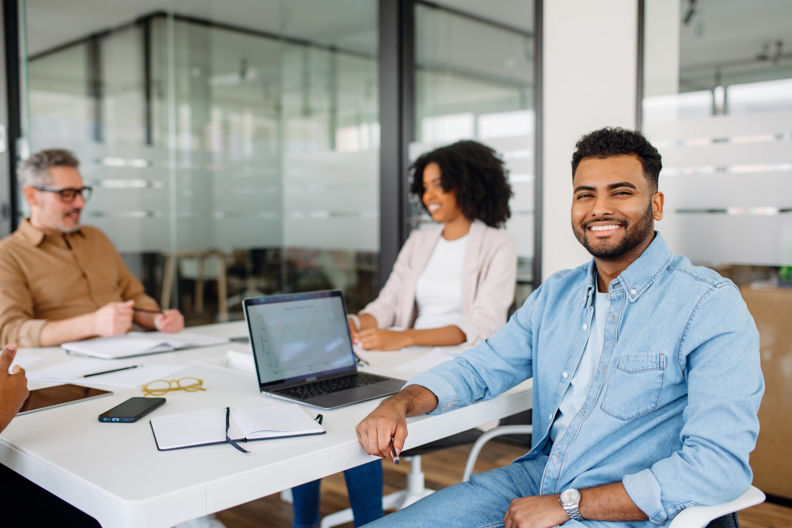 A confident young professional in a denim shirt smiling at the camera while sitting in a bright and modern office space. His positive energy reflects teamwork and a friendly work environment.