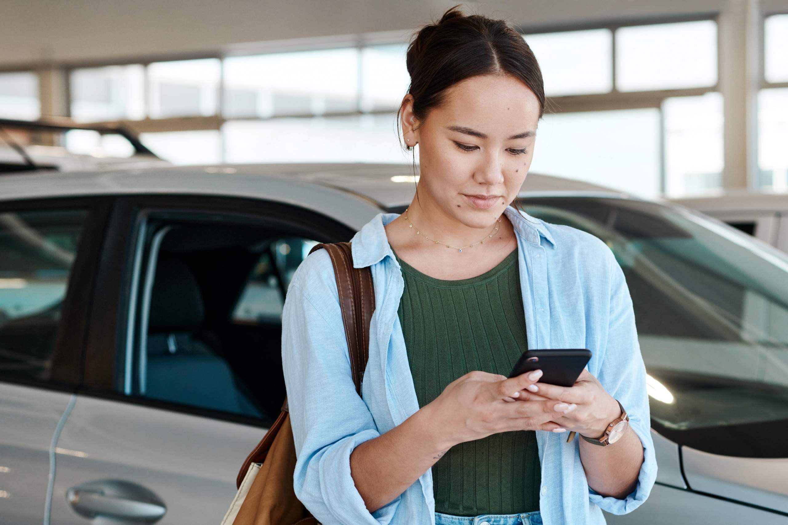 Asian woman, typing and phone at car dealership to sale, customer and website for research. Female person, communication and mobile app for vehicle review or online survey, showroom and social media
