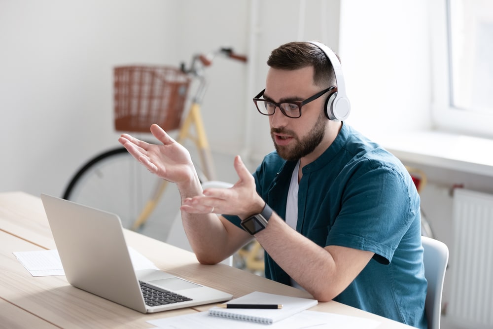 Focused young businessman in eyewear wearing headphones, holding video call with clients on laptop. Concentrated millennial man in glasses giving online educational class lecture, consulting customer.