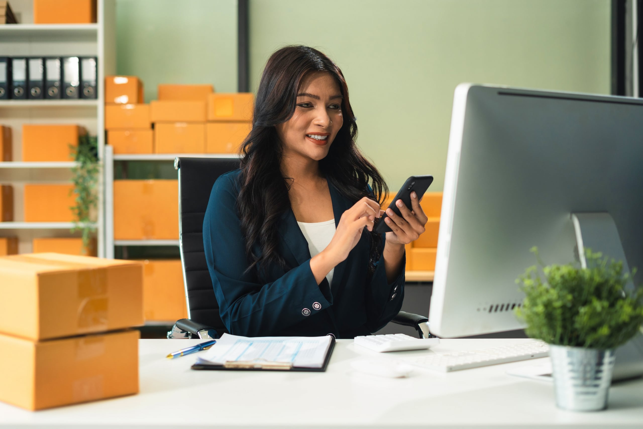 women smiling while on her cell phone while sitting at her computer with boxes and packages behind her
