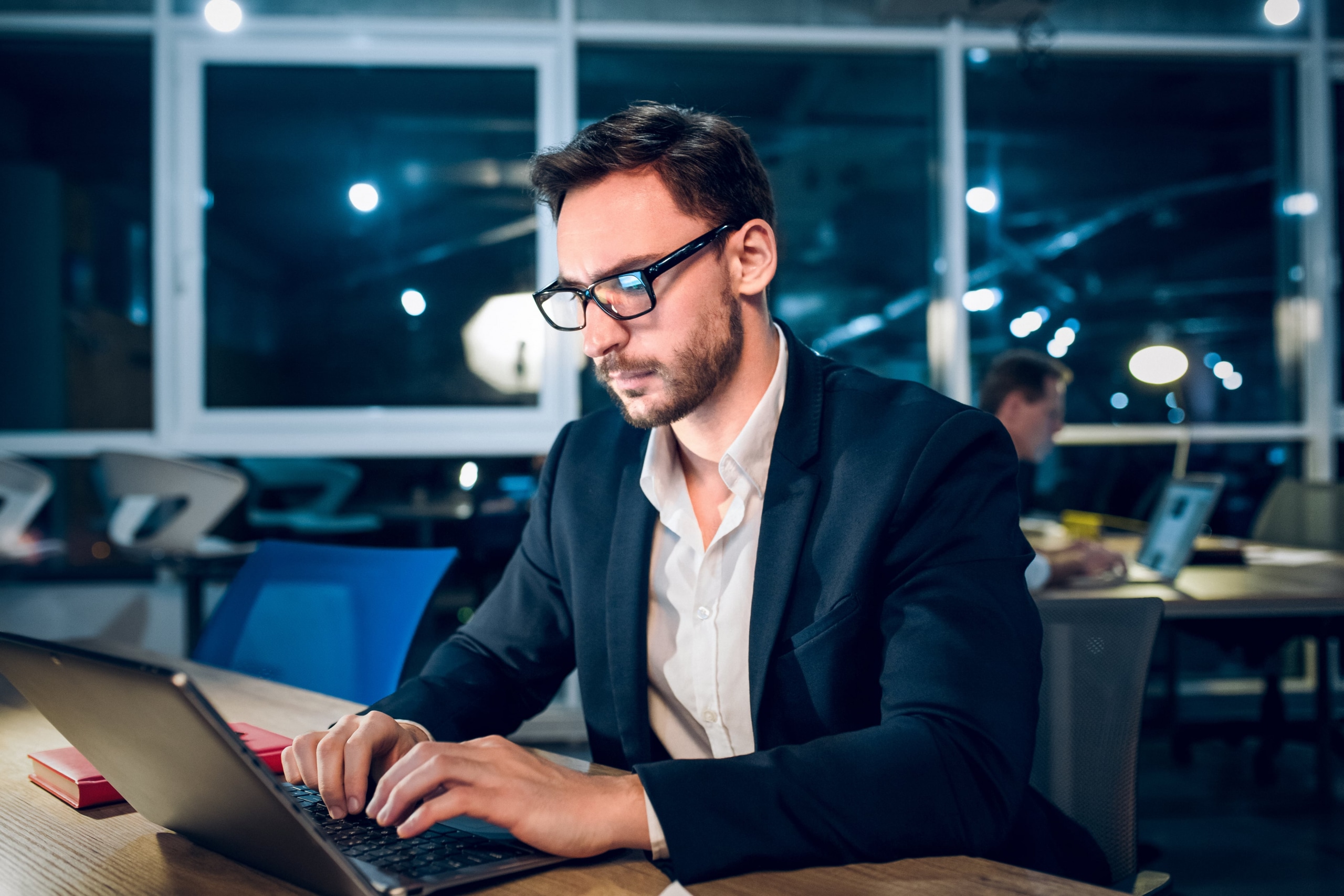 Bearded man with glasses typing on his laptop in a modern office at night