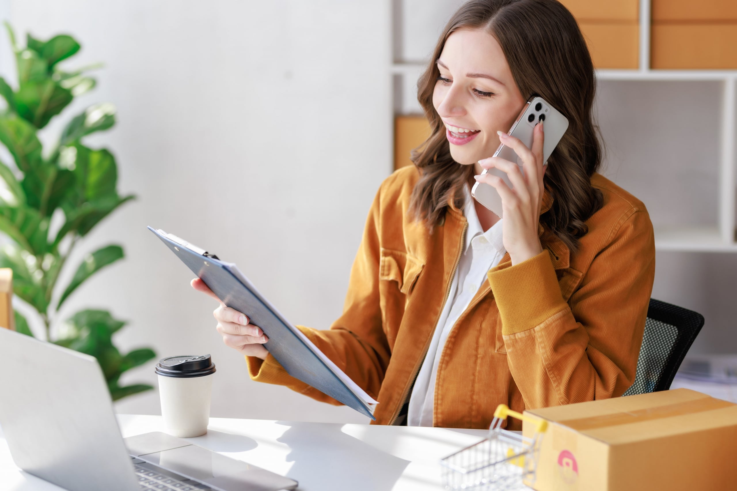 Young woman at her desk talking on her cell phone while looking at a clipboard