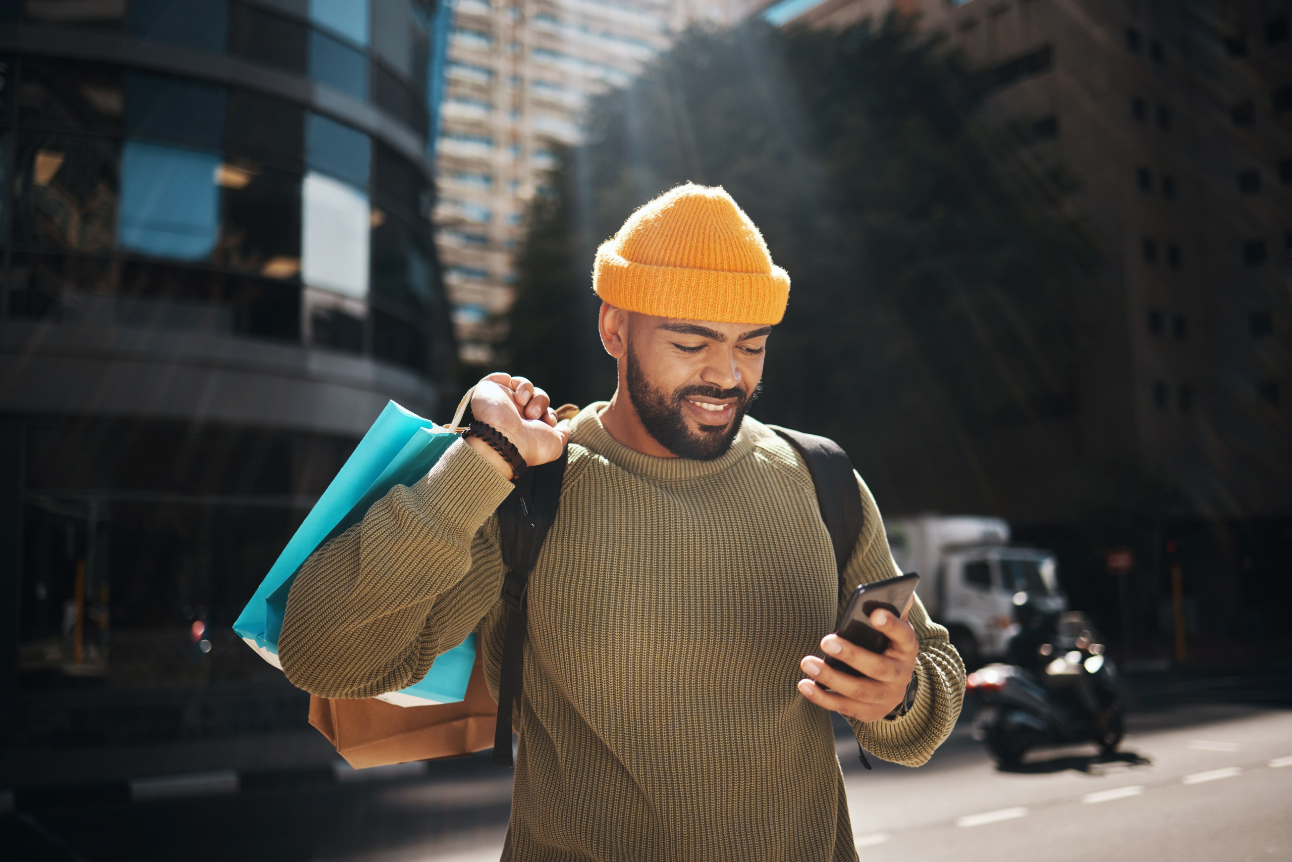 Man in a city holding shopping bags smiling looking down at his cell phone