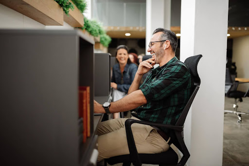 older man sitting at a cubicle with earbuds laughing with co worker