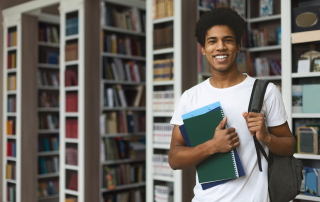 young african american college student in a library holding a book and his backpack