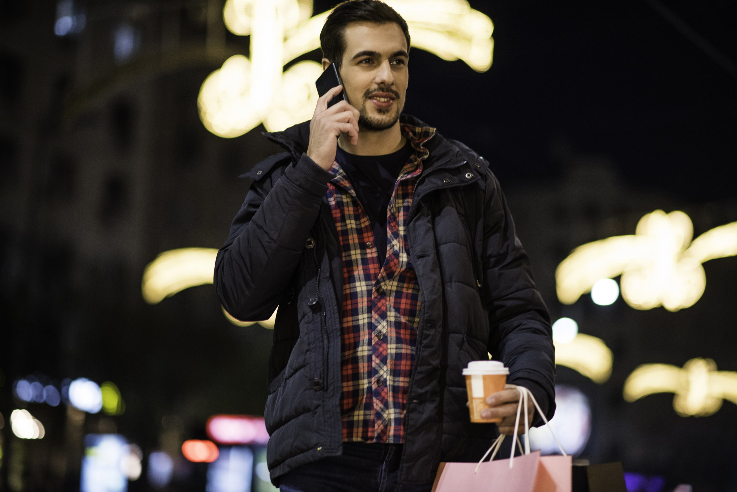 Bearded man talking on his phone at night in a city while holding a shopping bag